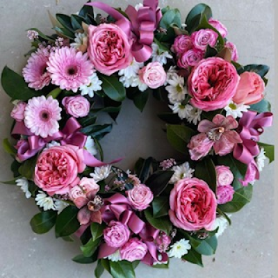 Rosy Radiance Wreath - The Rosy Radiance Wreath bursts with vibrant charm and heartfelt sentiment. Featuring a lush mix of bold pink garden roses, delicate daisies, gerberas, spray roses, and playful filler blooms, this wreath is accented with rich greenery and luxurious satin ribbons for a touch of elegance. Ideal for celebrations of life, special events, or as a vibrant tribute of love, this floral design radiates warmth, joy, and tender remembrance.